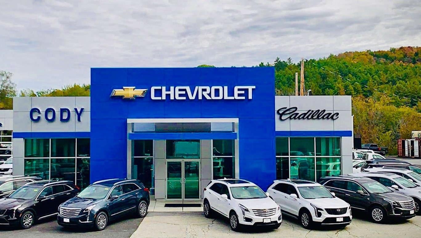 Cody Chevrolet Cadillac dealership building with multiple vehicles displayed in front and a wooded hill in the background.